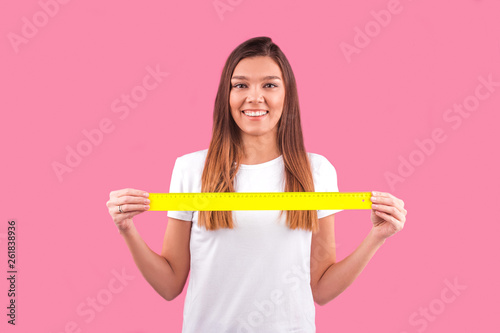 a young girl holds a plastic ruler on a pink background in front of her chest