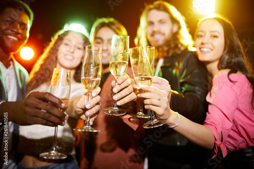 Close-up of young people in casual clothing standing in row and raising flutes while drinking champagne at party in nightclub