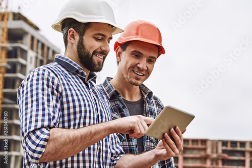 Working in team. Two young and cheerful builders in protective helmets are using digital tablet and working while standing at construction site