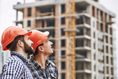 Teamwork. Two young and cheerful builders in red helmets are looking up and working at construction site photo