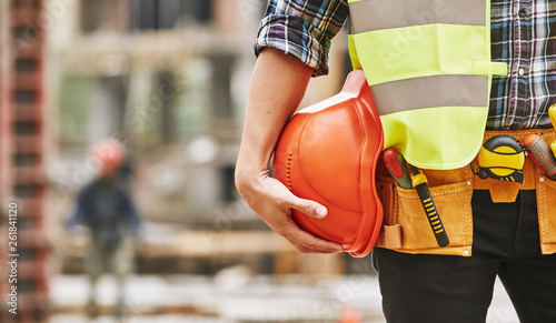 Construction worker. Cropped photo of male professional builder in working uniform with construction tools holding a safety red helmet while standing outdoor of construction site photo