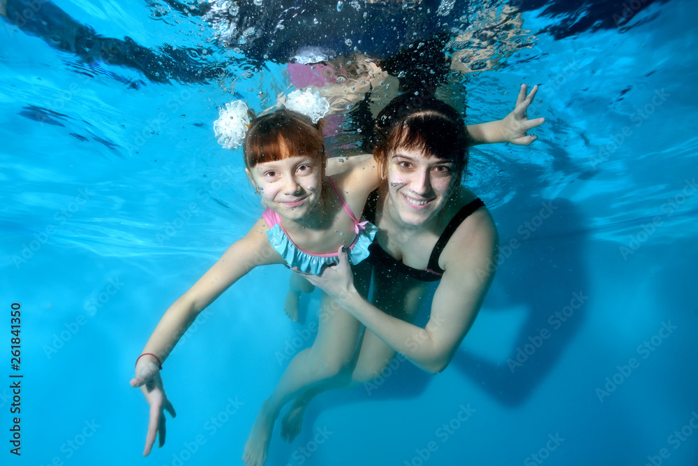 Happy mom and her little daughter play sports and swim underwater in the pool with their eyes open. They hug and smile under the water on a blue background. Portrait. Horizontal view