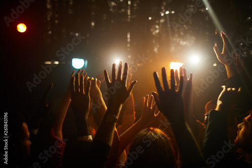 Crowd of unrecognizable people standing in dark club and waving hands in air while waiting for performer on stage