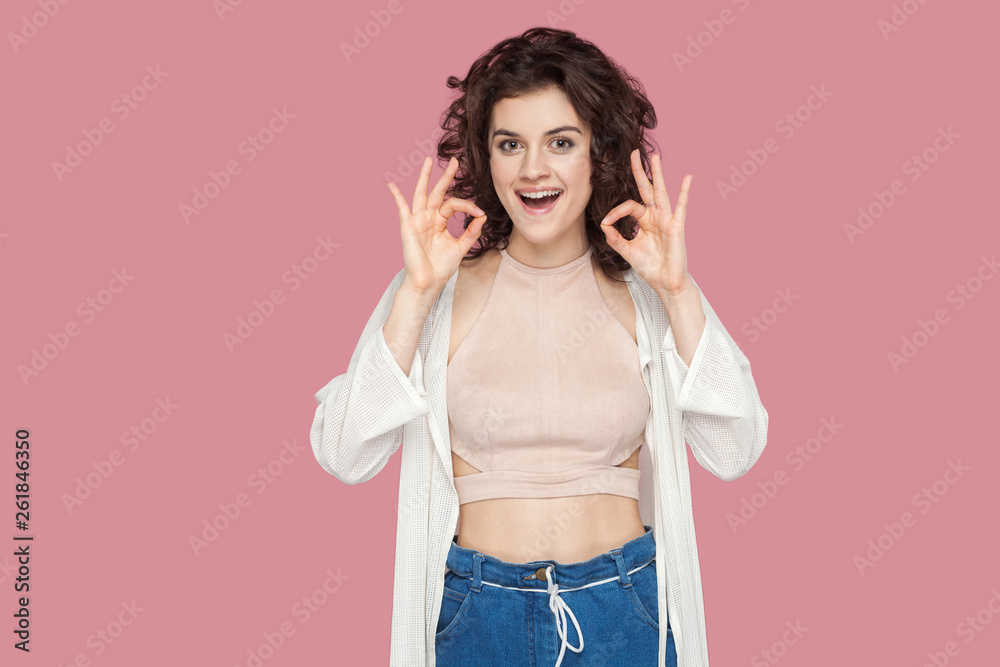 Portrait of funny excited beautiful brunette young woman with curly hairstyle in casual style standing with Ok or peace sign and looking at camera. indoor studio shot isolated on pink background.