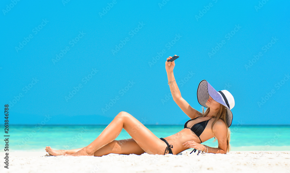 Cheerful young woman having fun taking smartphone selfie pictures of herself on tropical beach travel Caribbean holidays. Happy European girl model wearing fashion hat near sea. Summer lifestyle.