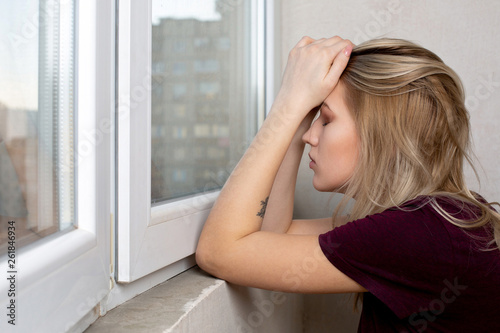 Sad blonde woman covering her head by hands sitting near the window. Space for text