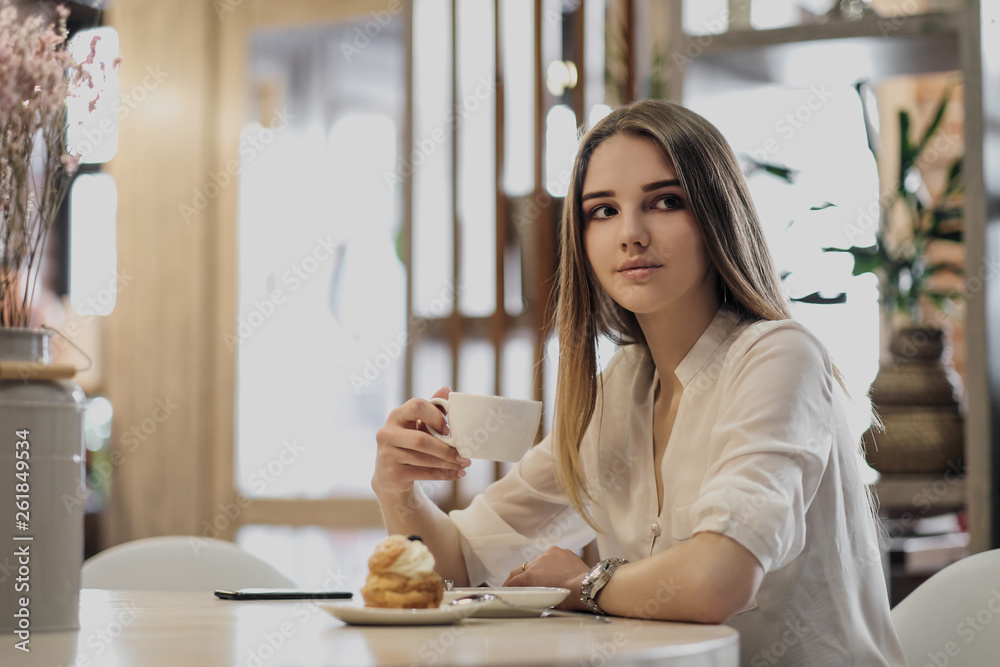 Young beautiful brown-haired girl in a white shirt is sitting at a table in a cafe by the window, drinking cappuccino coffee and dessert with whipped cream. Young beautiful brown-haired girl in a white shirt is sitting at a table in a cafe by the window, drinking cappuccino coffee and dessert with whipped cream.