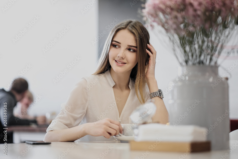 Young beautiful brown-haired girl in a white shirt is sitting at a table in a cafe by the window, drinking cappuccino coffee and dessert with whipped cream. Young beautiful brown-haired girl in a white shirt is sitting at a table in a cafe by the window, drinking cappuccino coffee and dessert with whipped cream.