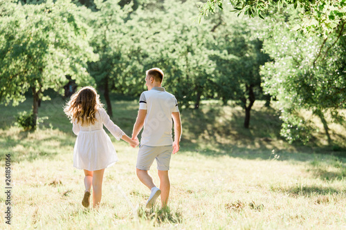 Back view of young pretty couple in love walking in park. Handsome cheerful blonde girl in white dress hugging her boyfriend. Man and woman having fun outdoors