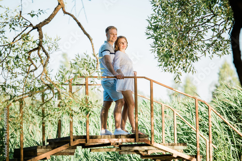 Young pretty couple in love standing on little bridge in park. Handsome cheerful blonde girl in white dress hugging her boyfriend. Man and woman having fun outdoors