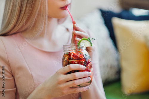 Young beautiful blonde girl in a pink dress sits at a cafe table, drinks lemonade from a glass jar.
