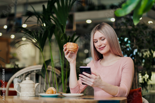 Young beautiful girl sitting in a green cafe at a table. Drinking tea with croissants, chatting, laughing and taking pictures on a smartphone.