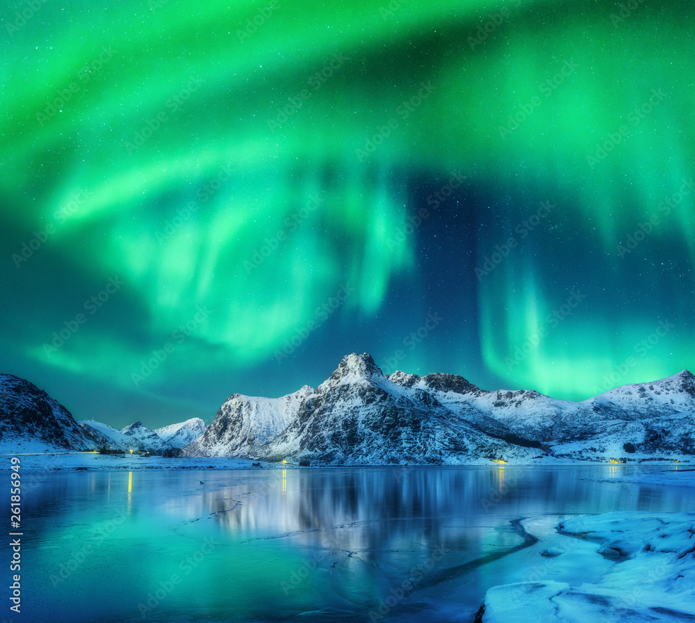 Aurora borealis over snowy mountains, frozen sea coast and reflection in water in Lofoten islands, Norway. Northern lights. Winter landscape with polar lights, ice in water. Starry sky with aurora