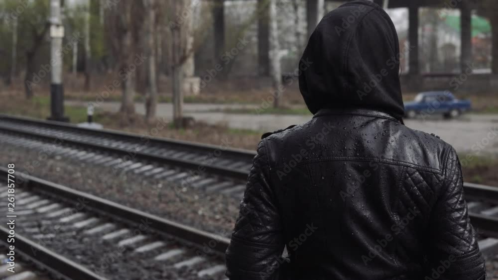 A young woman stands on the platform and waiting for the train. Female in a leather jacket with a hood, looking away on a rainy day. A young woman stands on the platform and waiting for the train. Female in a leather jacket with a hood, looking away on a rainy day.