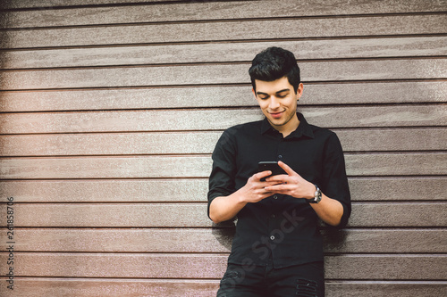 Portrait of young handsome Turkish male model. Mediterranean race brunette in black shirt is using a hand phone of a technology businessman on the street in sunny weather on a wooden wall background