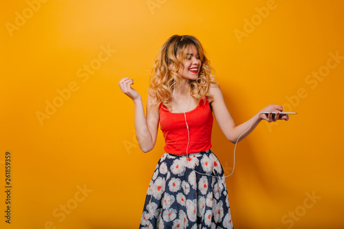Pretty girl with long curly hair in tail listening to music with headphones on orange background in studio. She wears red T-shirt, skirt flowers, red lips. She is dancing. She looks excited.