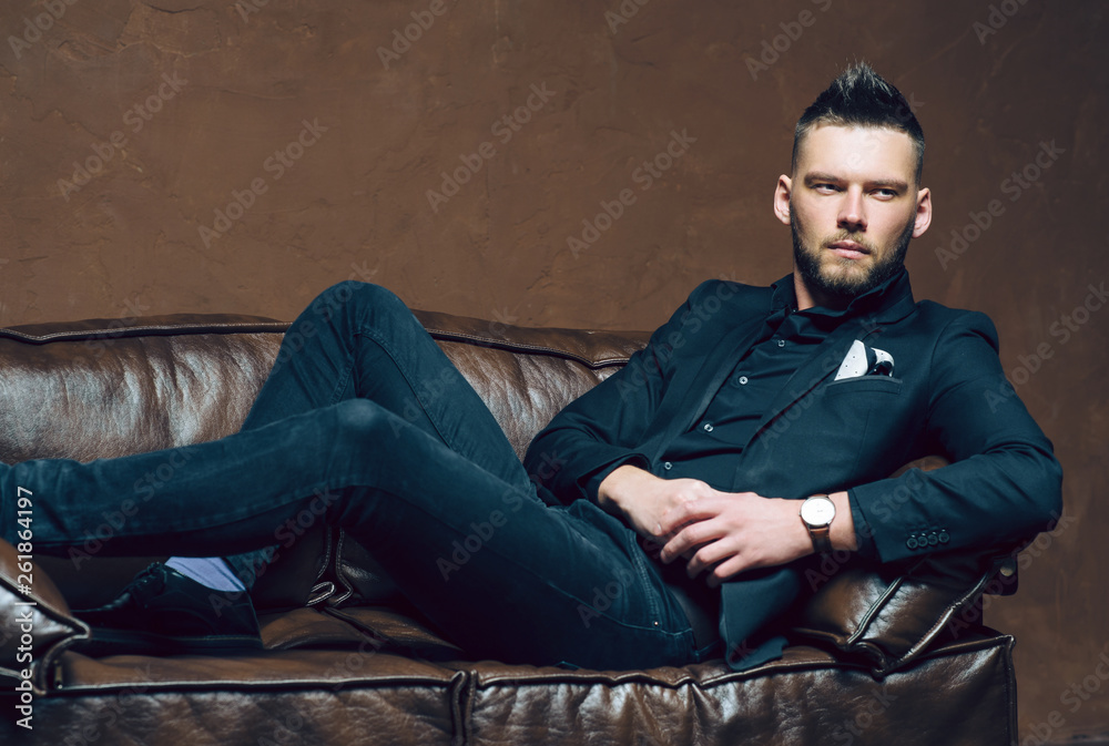 Young handsome man posing for a fashion shoot in a studio. Fashion as a lifestyle. Man wearing a beard. Model sitting on a sofa in tuxedo. Successful fashionable businessman. Business look advertising Young handsome man posing for a fashion shoot in a studio. Fashion as a lifestyle. Man wearing a beard. Model sitting on a sofa in tuxedo. Successful fashionable businessman. Business look advertising