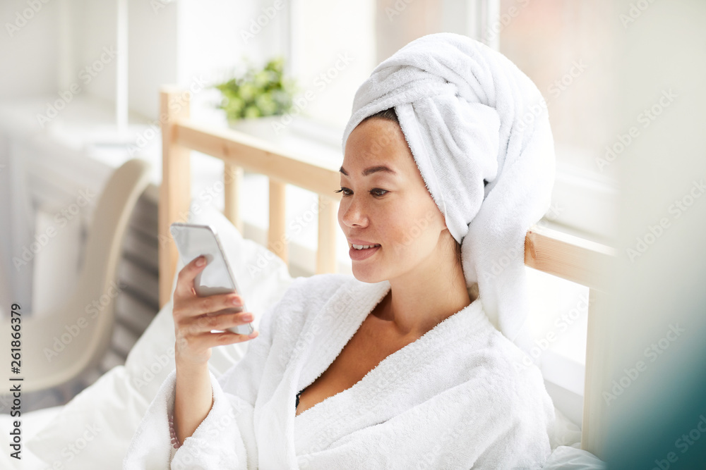 Side view portrait of smiling Asian woman wearing bath robe using smartphone while relaxing in bed at home, copy space Side view portrait of smiling Asian woman wearing bath robe using smartphone while relaxing in bed at home, copy space