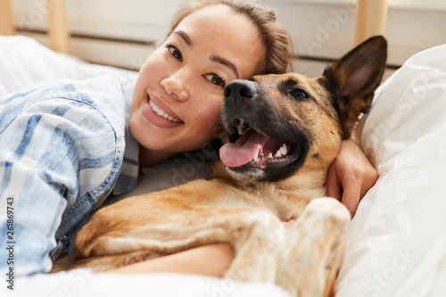 Portrait of smiling Asian woman hugging dog lying on bed together and looking at camera, copy space