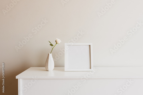 Close up of single rose in small white vase next to blank square picture frame on sideboard against neutral wall - warm matte filter effect photo