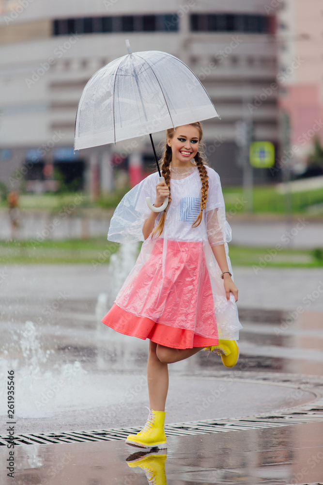 Young pretty girl with two braids in yellow boots and with transparent umbrella stands near fountain.