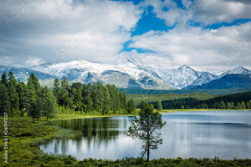 Small lake on grass and fluffy clouds over green meadows and snowy peaks. Highland lake  Altay  Siberia.