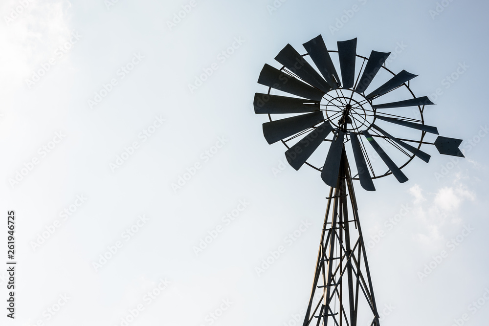 Low angle view of an old-fashioned, multi-bladed, metal wind pump atop a lattice tower in backlight against a pale blue sky. Low angle view of an old-fashioned, multi-bladed, metal wind pump atop a lattice tower in backlight against a pale blue sky.