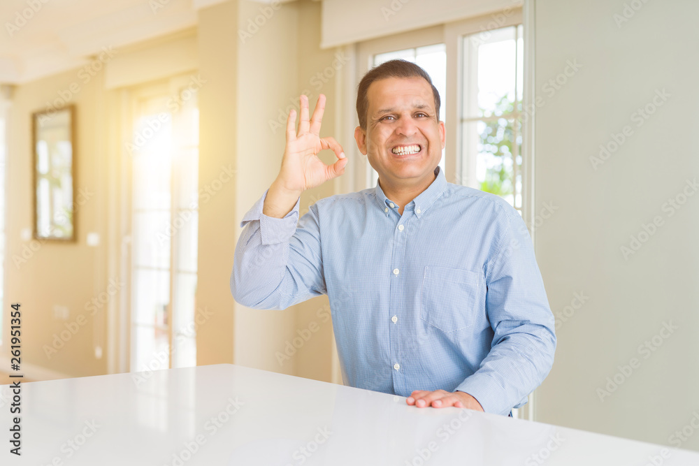 Middle age man sitting at home smiling positive doing ok sign with hand and fingers. Successful expression.