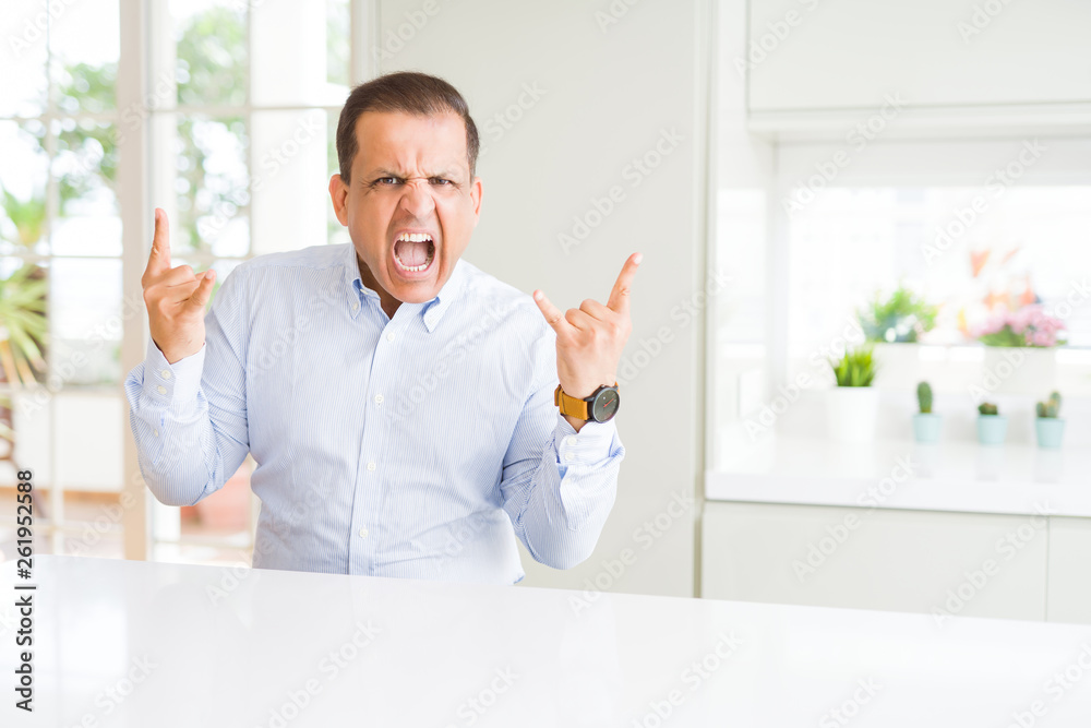 Middle age man sitting at home shouting with crazy expression doing rock symbol with hands up. Music star. Heavy concept.