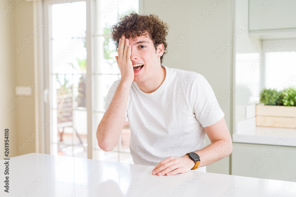 Young handsome man wearing white t-shirt at home covering one eye with hand with confident smile on face and surprise emotion.