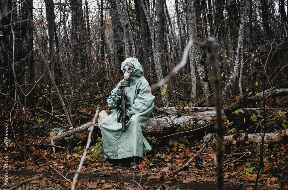 Stalker, a man in a gas mask and special chemical protection, green cloak.  With a gun in his hand, he sits on a stump in the forest.