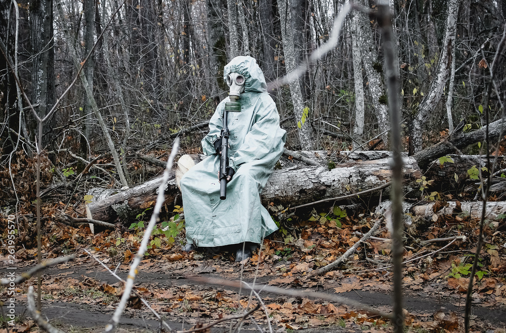 Stalker, a man in a gas mask and special chemical protection, green cloak.  With a gun in his hand, he sits on a stump in the forest.