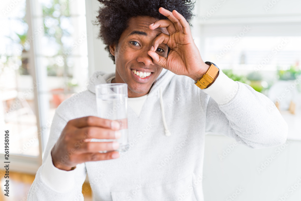 African American man drinking a glass of water at home with happy face smiling doing ok sign with hand on eye looking through fingers