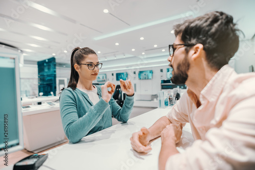 Young Caucasian charming brunette with ponytail and eyeglasses dressed casual explaining to seller which smart phone she want to buy. Tech store interior.
