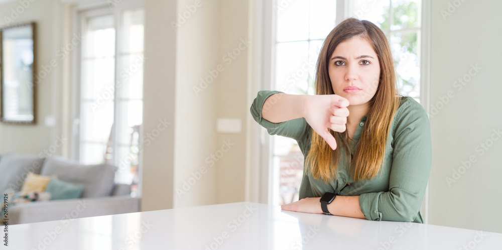 Beautiful young woman at home looking unhappy and angry showing rejection and negative with thumbs down gesture. Bad expression.