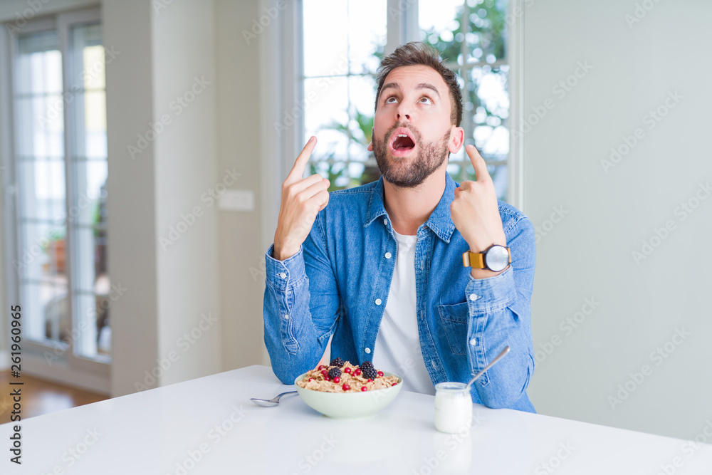 Handsome man eating cereals for breakfast at home amazed and surprised looking up and pointing with fingers and raised arms.