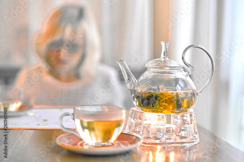 Transparent teapot with green tea on a stand with a candle on the background of a blurred breakfast blonde