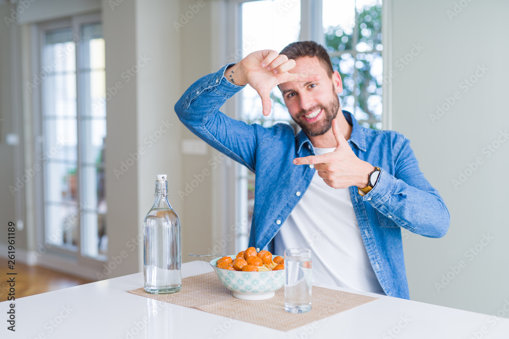 Handsome man eating pasta with meatballs and tomato sauce at home smiling making frame with hands and fingers with happy face. Creativity and photography concept.