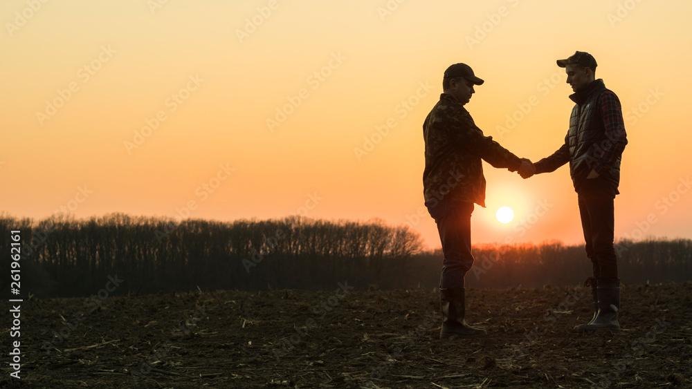 Two male farmers shake hands with each other. Stand on the background of a plowed field at sunset