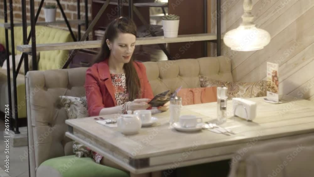 Beautiful European elegant young woman girl in red coral jacket reading the menu booklet in a cozy glamorous cafe Beautiful European elegant young woman girl in red coral jacket reading the menu booklet in a cozy glamorous cafe