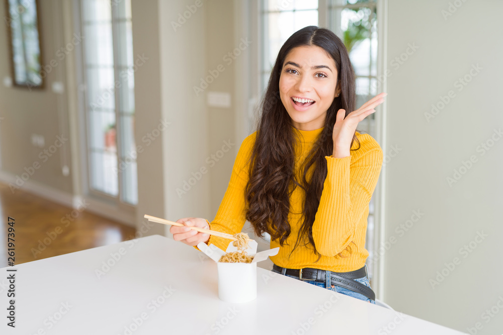 Young woman eating noodles in delivery box very happy and excited, winner expression celebrating victory screaming with big smile and raised hands Young woman eating noodles in delivery box very happy and excited, winner expression celebrating victory screaming with big smile and raised hands