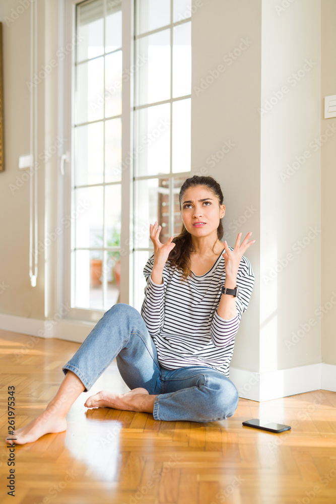 Young beautiful woman sitting on the floor at home crazy and mad shouting and yelling with aggressive expression and arms raised. Frustration concept. Young beautiful woman sitting on the floor at home crazy and mad shouting and yelling with aggressive expression and arms raised. Frustration concept.