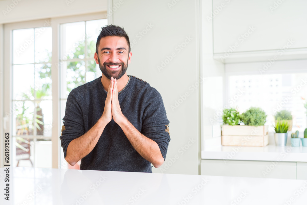 Handsome hispanic man wearing casual sweater at home praying with hands together asking for forgiveness smiling confident.