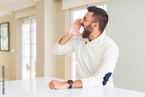 Handsome hispanic man wearing casual white sweater at home shouting and screaming loud to side with hand on mouth. Communication concept.