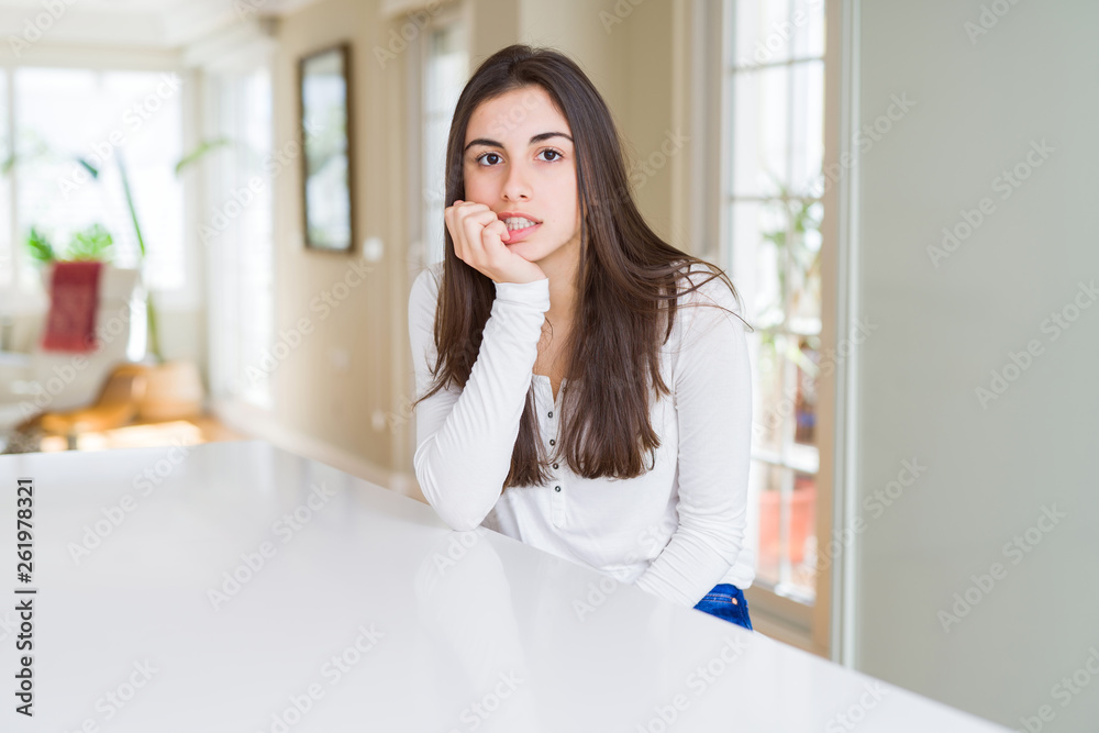 Beautiful young woman sitting on white table at home looking stressed and nervous with hands on mouth biting nails. Anxiety problem. Beautiful young woman sitting on white table at home looking stressed and nervous with hands on mouth biting nails. Anxiety problem.