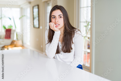 Free Download Beautiful young woman sitting on white table at home looking stressed and nervous with hands on mouth biting nails. Anxiety problem. - GFXLoad