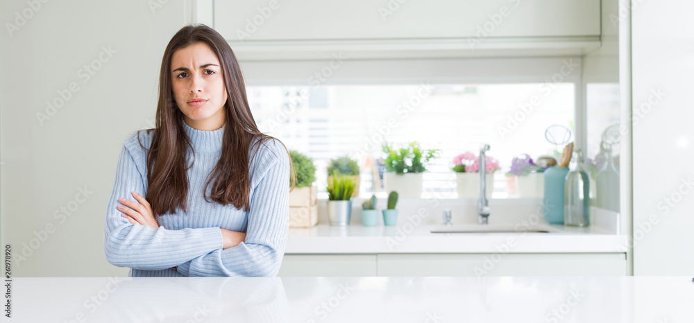 Wide angle picture of beautiful young woman sitting on white table at home skeptic and nervous, disapproving expression on face with crossed arms. Negative person. Wide angle picture of beautiful young woman sitting on white table at home skeptic and nervous, disapproving expression on face with crossed arms. Negative person.