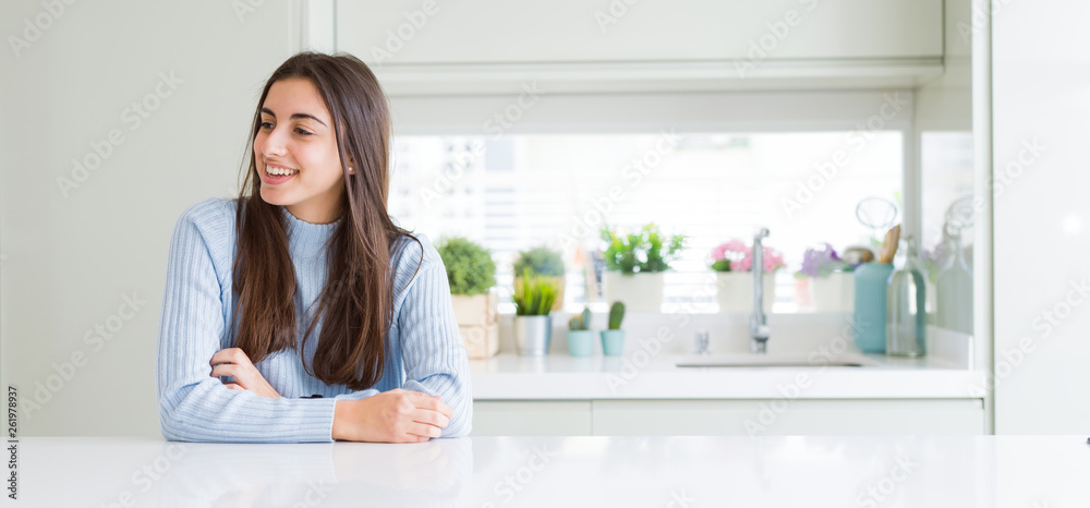 Wide angle picture of beautiful young woman sitting on white table at home looking away to side with smile on face, natural expression. Laughing confident. Wide angle picture of beautiful young woman sitting on white table at home looking away to side with smile on face, natural expression. Laughing confident.