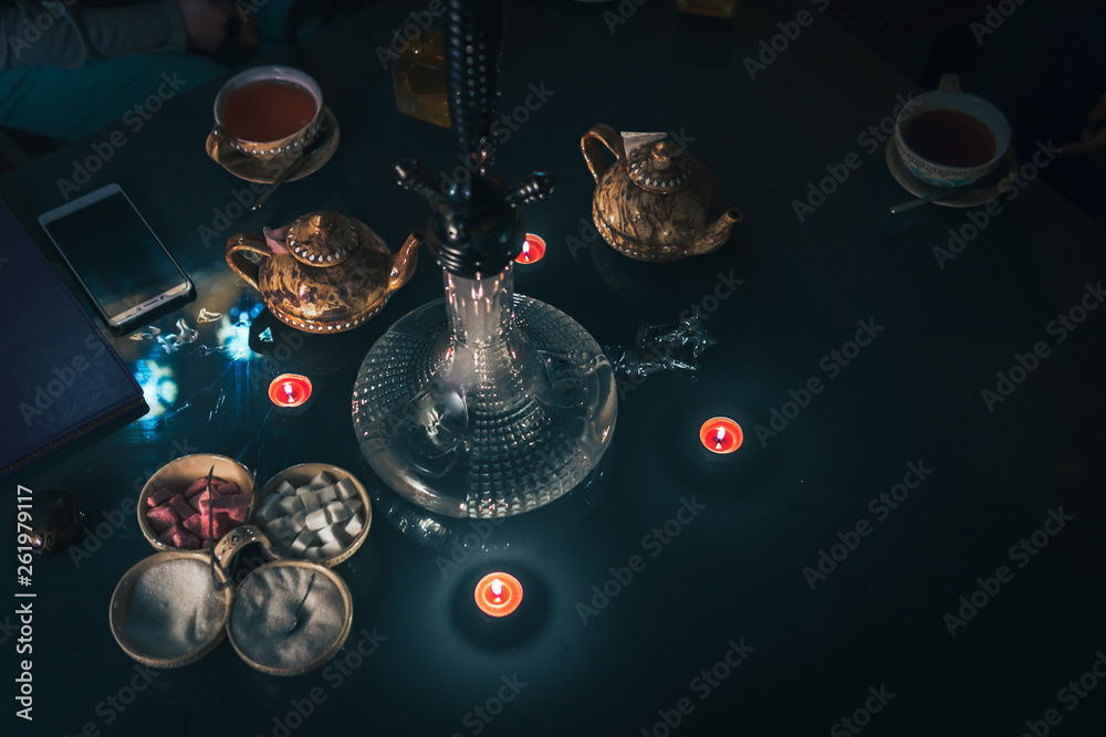 Hookah on glass table top view. cups candle and smartphone eastern tea ceremony. Stylish oriental shisha in dark with backlight. Selective focus, sugar cubes with different flavors. Copy space