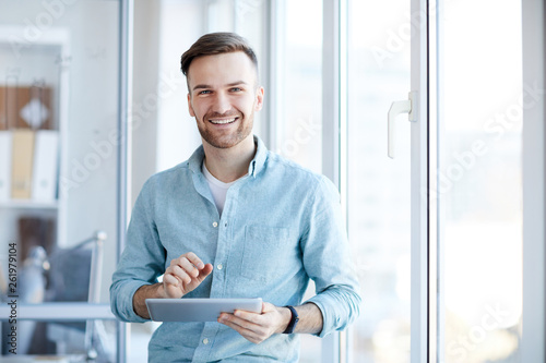 Waist up portrait of handsome young man holding digital tablet and smiling happily at camera standing by window, copy space